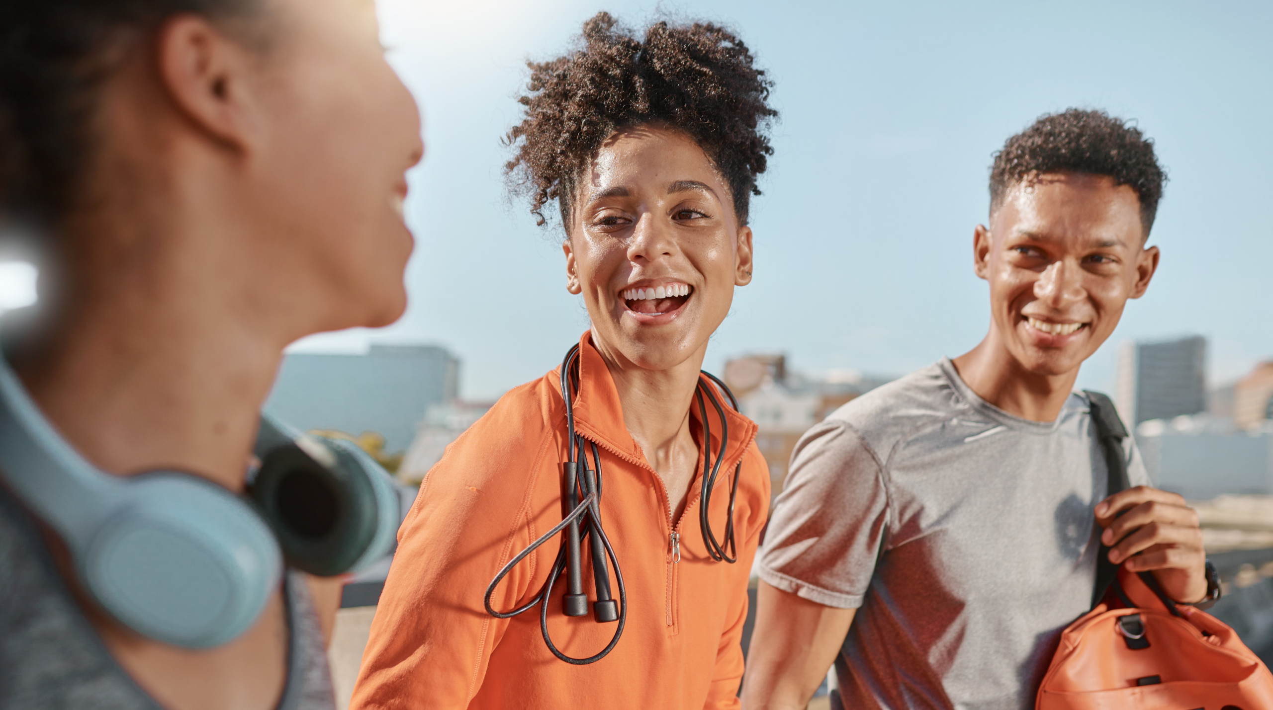 Three friends in workout clothes smile and chat outdoors, with one person wearing a stethoscope and city buildings blurred in background.